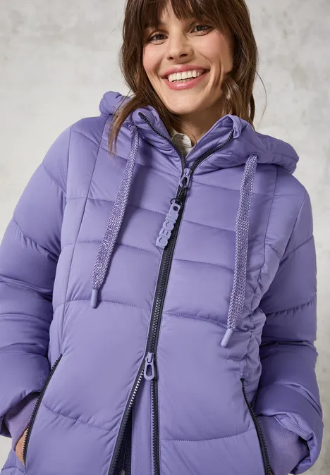 Frau mit lila Steppjacke und Kapuze lächelt.